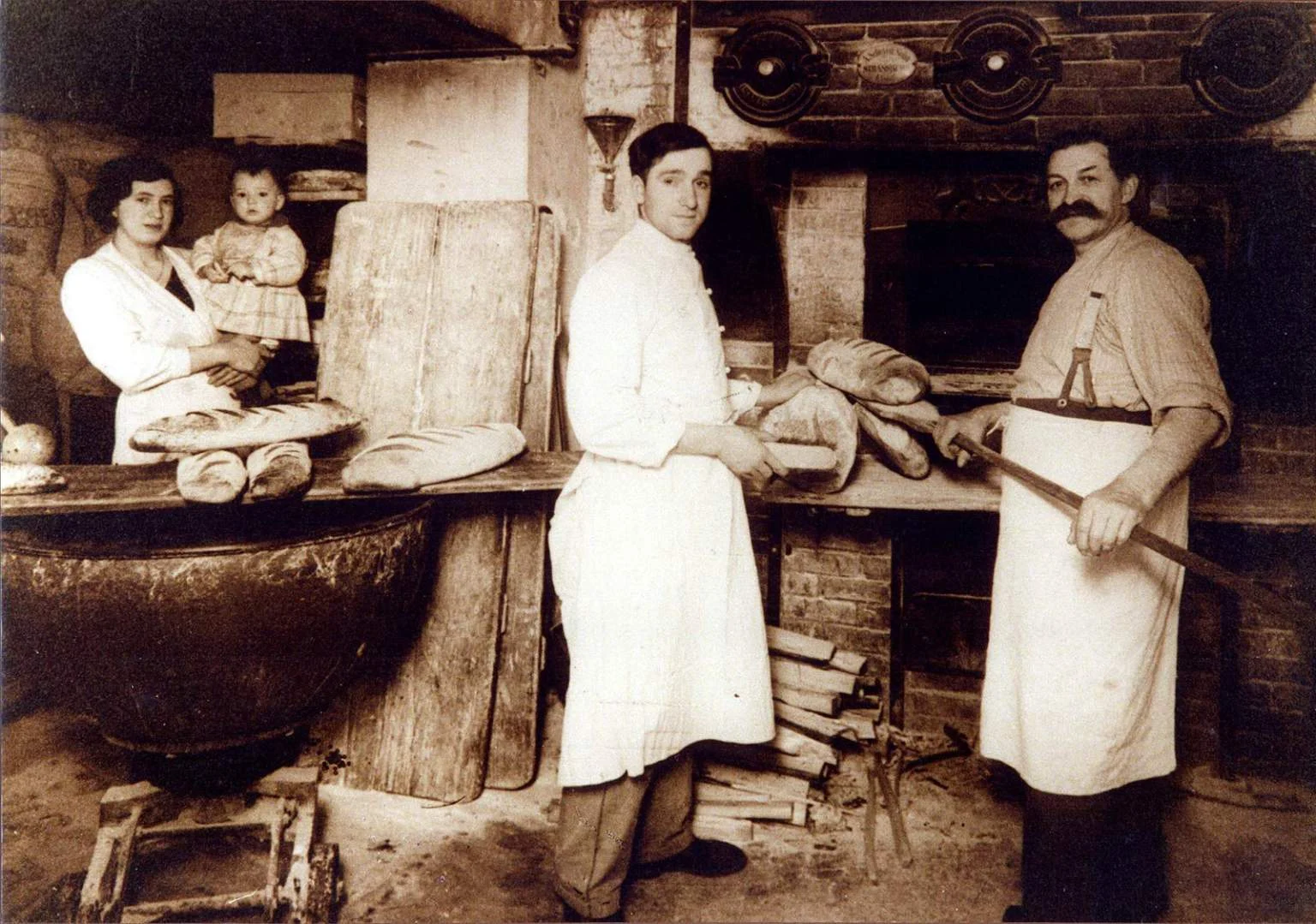 Hotel-Restaurant-Entzheim-Strasbourg-Pere-Benoit-Histoire Vieille photo de la cuisine du Restaurant Steinkeller à Entzheim : 2 chefs cuisiniers autour d'un vieux four à pain et une femme tenant un enfant dans ses bras